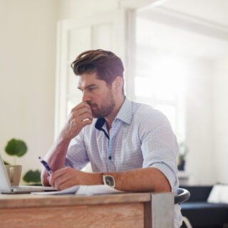 Man working on a laptop in a home office, seated at a desk with a focused expression. The workspace is bright and organised, creating a professional remote working environment.